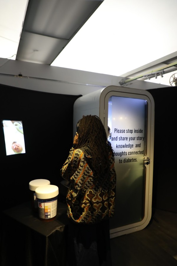 An exhibit visitor wearing headphones watches a featured video while standing next to two oversized pill bottles on the left side and a private sound-proof booth for sharing feedback on the right. On the door of the booth, it reads “Please step inside and share your story, knowledge and thoughts connected to diabetes.”